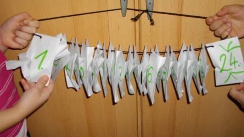 Zwei Hände halten einen selbstgemachten Adventskalender mit nummerierten Papiertüten von 1 bis 24 an einem Faden vor einem Holzschrank.