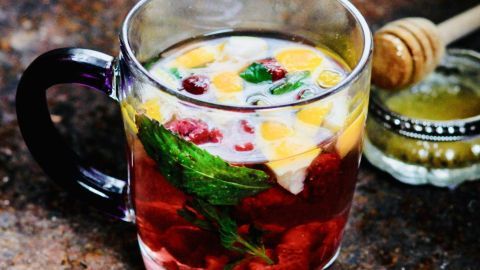 Ein durchsichtiges Glas mit erkältungstee, gefüllt mit Ingwer, Früchten und Minze, steht auf einer rustikalen Oberfläche.