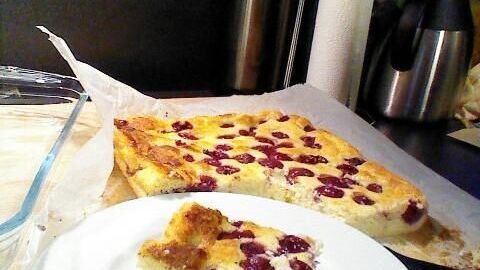 Ein Stück Käsekuchen mit Kirschen liegt auf einem weißen Teller, während der restliche Kuchen im Hintergrund auf Backpapier ruht.