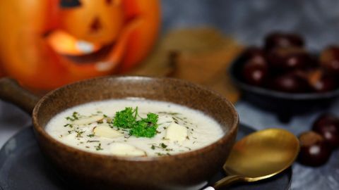 Die Kastaniencremesuppe wird in einer Schüssel serviert, dekoriert mit Petersilie, während im Hintergrund eine leuchtende Kürbisdekoration ist.