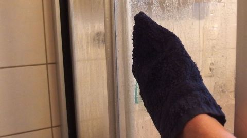 Ein Handtuch reibt über eine beschlagene Duschwand, um Kalk zu entfernen und die Fliesen im Badezimmer zu reinigen.