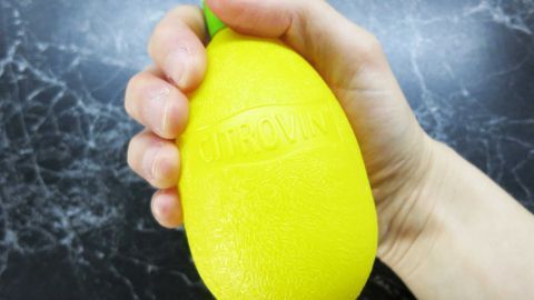 Zitronensaft für strahlend weiße Nägel: Reibe deine verfärbten Nägel regelmäßig mit Zitronensaft ein und entdecke die natürliche Kraft der Zitrone für einen klaren, frischen Look. Einfach und effektiv! Eine Hand hält eine gelbe Plastikzitrone mit der Aufschrift "CITROVIN" vor einem dunklen Marmortisch.