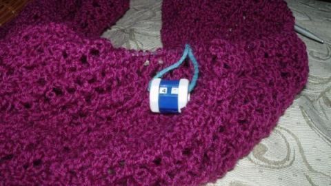 Ein Reihenzähler liegt auf einer lila Strickware, während das Stricken in einem gemütlichen Raum stattfindet.