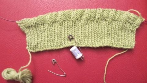 Ein gestricktes grünes Stück liegt auf rotem Untergrund mit zwei Sicherheitsnadeln und einem Reihenzähler.