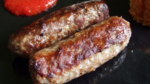 Zwei gebratene Cevapcici liegen auf einem dunklen Teller, neben einer Portion roter Sauce.