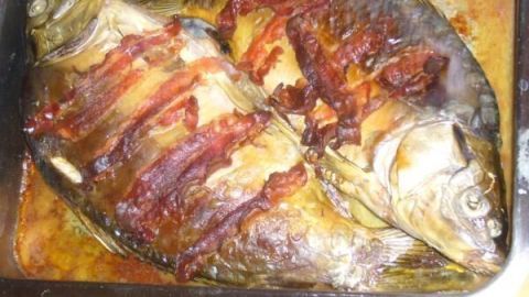 Speck-Karpfen liegt gebraten in einer Ofenform, bedeckt mit knusprigem Speck, umgeben von Bratensaft auf einem Tisch.