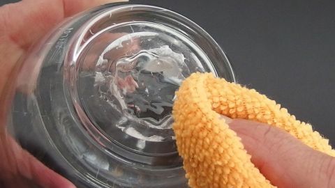 Eine Hand hält ein Glas mit Aufkleberresten, während ein gelbes Tuch über die Oberfläche zum Entfernen der Rückstände wischt.