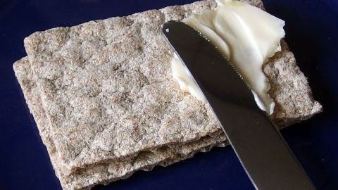 Knäckebrot liegt auf einem blauen Teller, während ein Messer darauf Butter verteilt, um es zu belegen.