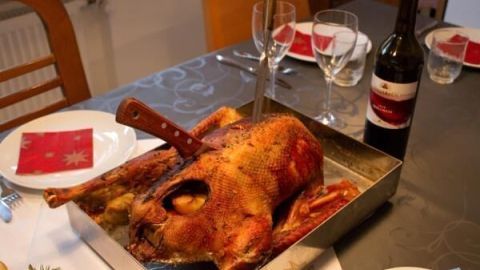 Gänsebraten liegt mit einem Messer in einer Pfanne auf einem festlich gedeckten Tisch; ein Rotwein steht daneben.