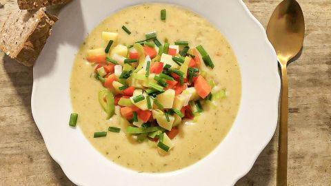 Eine Schale Samtsuppe mit bunten Gemüsestückchen liegt auf einem Holztisch, begleitet von frischem Brot und einem Löffel.