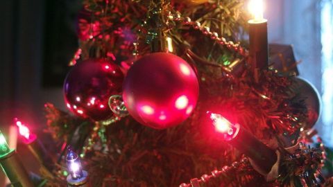 Ein Weihnachtsbaum wird festlich mit einer bunten Lichterkette und ornamentalen Kugeln geschmückt, umgeben von warmem Licht.