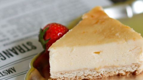 Ein Stück Cheesecake deluxe liegt auf einem Teller, dekoriert mit einer Erdbeere, während ein Rezeptblatt im Hintergrund sichtbar ist.