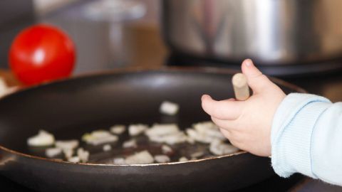 Ein kleines Kind greift mit einer Hand nach einem Kochlöffel in einer Pfanne, während Zwiebeln darin braten.
