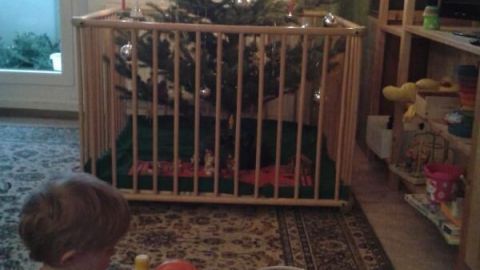 Ein Krabbelkind sitzt vor einem Weihnachtsbaum und spielt mit einem Spielzeug, während der Raum festlich dekoriert ist.