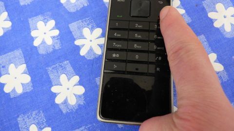 Ein Finger drückt eine Taste eines Telefons auf einem blauen Tischtuch mit weißen Blumen, um Werbeanrufe abzuwehren.