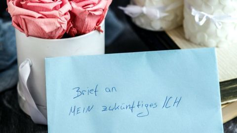 Ein Brief in einem blauen Umschlag liegt vor einem Kerzenarrangement und einer rosa Blütenbox mit der Aufschrift „Brief an mein zukünftiges Ich“.