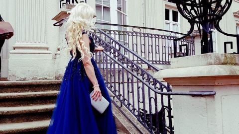 Eine Frau in einem langen, blauen Abendkleid geht elegant eine Treppe zu einem prächtigen Gebäude hinauf.