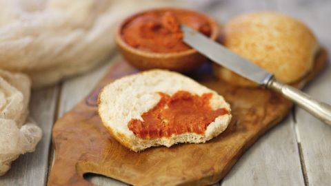 Eine belegte Brötchenhälfte mit Tomatenbutter liegt auf einem Holzbrett, neben einer Schüssel mit der restlichen Butter.