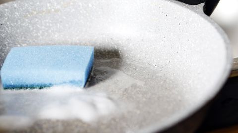 Eine stark verschmutzte Keramikpfanne liegt im Spülbecken mit einem blauen Schwamm und Seife zum Reinigen.
