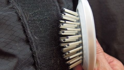 Eine Hand hält eine Bürste, die Fussel von einem Klettverschluss auf einem schwarzen Stoff entfernt.