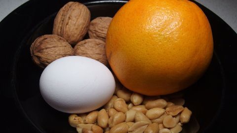 Eine Schüssel mit Nüssen, einer Orange, einem Ei und Erdnüssen steht auf einem Tisch, ideal für gesundes Zunehmen.
