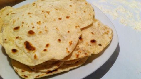 Auf einem Teller liegen mehrere Chapatis, die gleichmäßig gebräunt sind. Im Hintergrund sind Küchenutensilien und Mehl zu sehen.