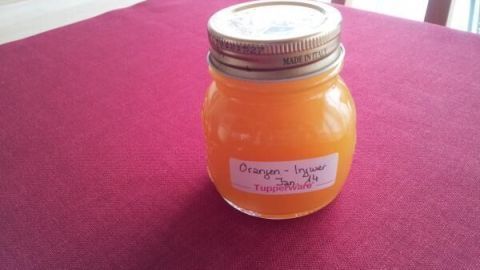 Ein Glas Orangen-Ingwer-Konfitüre steht auf einem roten Tisch. Das leuchtend orangefarbene Produkt ist in einem Schraubglas abgefüllt.