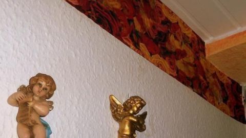 Engel aus Porzellan schmücken die Wand unter einer Wandbordüre mit leuchtendem Rosenmuster, neben einem roten Spiegel.