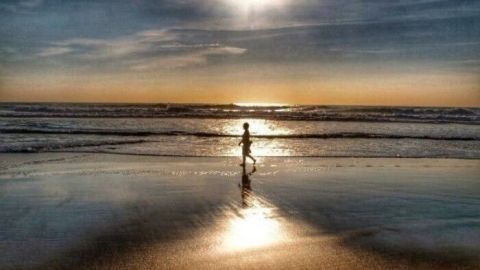 Ein Kind steht am Strand und spielt im Wasser, während die Sonne am Horizont untergeht und den Himmel erleuchtet.
