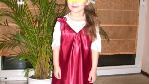 Ein Kind trägt ein Dornröschen-Faschingskostüm: ein langes rotes Kleid und eine Blumenkronen, steht vor Pflanzen im Wohnzimmer.