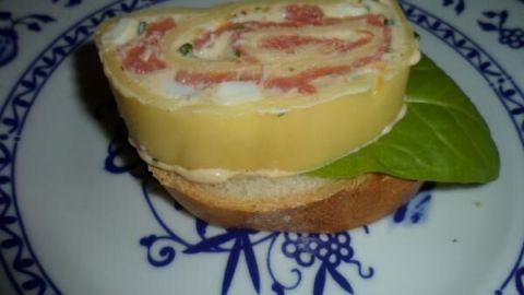 Eine Käse-Lachs-Rolle liegt auf einem Teller mit einem Stück Brotes und einem Blatt frischem Grün, appetitlich angerichtet.