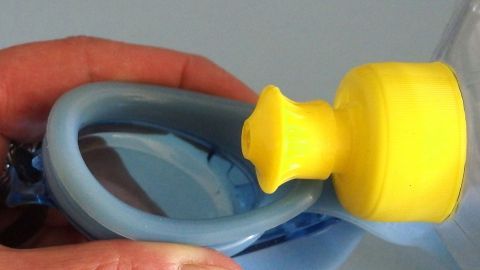 Eine Hand hält eine Schwimmbrille, während ein gelber Stopfen die Gläser vor dem Beschlagen schützt.