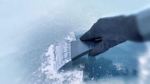 Eine behandschuhte Hand schabt mit einem Eiskratzer Eis von einer Autoscheibe; frostige Umgebung.