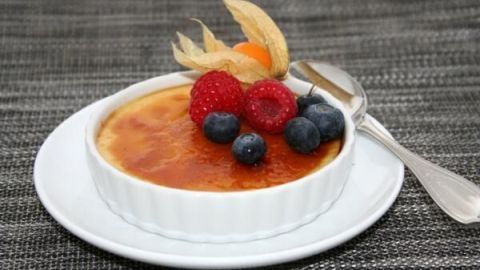 Crème brûlée in weißer Schale, garniert mit Beeren und Physalis, steht auf einem Tischset mit Löffel daneben.