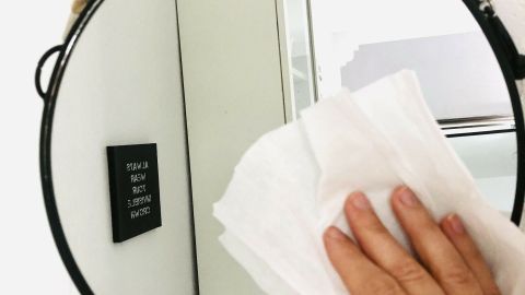 Eine Hand reinigt mit einem Babyfeuchttuch einen runden Spiegel, der an einer Wand hängt. Im Hintergrund steht ein Kühlschrank.