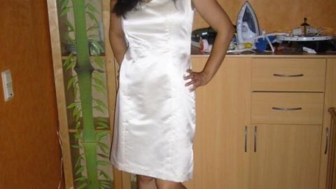 Eine Frau in einem eleganten weißen Kleid posiert lächelnd in einem wohnlichen Raum mit Holzschrank und Wandkunst.