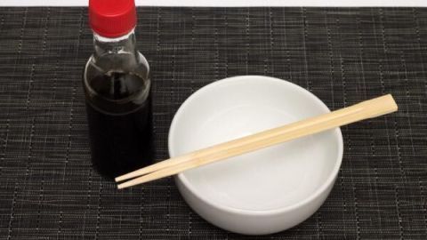 Eine Flasche Sojasauce steht neben einer leeren weißen Schale mit Essstäbchen auf einer schwarzen Matte.