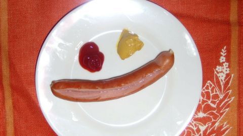 Ein Würstchen liegt auf einem weißen Teller, umgeben von Ketchup und Senf auf einem orangefarbenen Tisch.