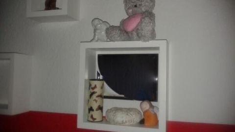 Ein Teddybär sitzt auf einem Regal, umgeben von weiteren Spielzeugen, in einem Raum mit rot-weißer Wandgestaltung.