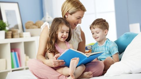 Eine Mutter liest mit ihren zwei Kindern in einem hellen Raum ein Buch; eine liebevolle, entspannte Atmosphäre fördert das Gespräch über Probleme.