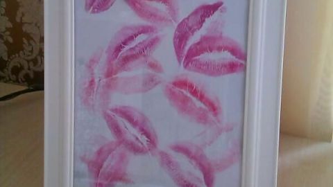 Gerahmtes Bild zeigt pinke Lippenstiftabdrücke in Weißrahmen, dekorativ auf Holztisch vor gemusterter Tapete.