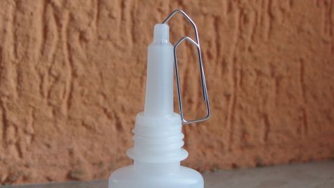 Eine Flasche Sekundenkleber steht vor einer strukturierten Wand, die Tülle wird durch einen Drahtverschluss freigehalten.