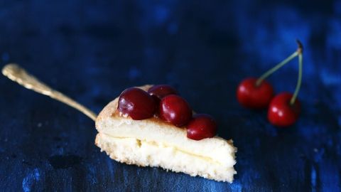 Ein Kirschfladenstück liegt auf einer silbernen Gabel, garniert mit frischen Kirschen, auf einem blauen Untergrund.