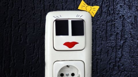 Ein dekorativer Lichtschalter in Form eines Gesichts mit roten Lippen und einem gelben Schleifchen, platziert an einer dunklen Wand.