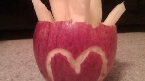 Apfelstückchen, geschnitten wie Pommes, stecken in einer roten Apfelhälfte mit M-Förmung, auf einer Küchenplatte.