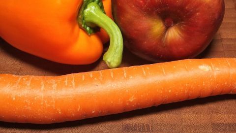 Eine orange Paprika, eine rote Apfel und eine Karotte liegen auf einem Holzbrett, ideal für Rohkost und FDH.
