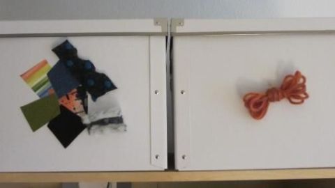 Zwei weiße Sortierboxen stehen nebeneinander; eine zeigt bunte Stoffstücke, die andere eine rote Kordel als Beschriftung.