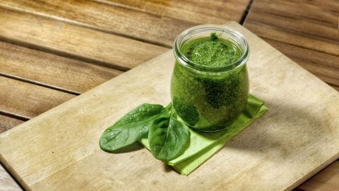 Ein Glas Pesto aus Blattsalaten steht auf einem Holzbrett, umgeben von frischen Spinatblättern und einer servierten Serviette.