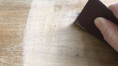 Eine Hand schmirgelt mit einem Schleifblock über eine helle Holzoberfläche, um weniger Staub zu erzeugen.
