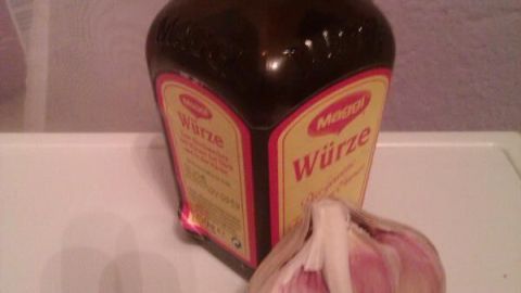 Ein Maggi Würze Glasflasche steht auf einem Tisch neben einer Knoblauchknolle vor einer lilafarbenen Wand.
